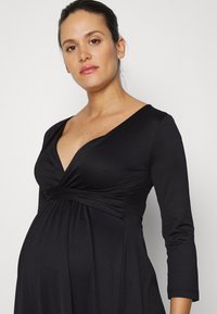 Vestido de maternidade preto com mangas longas e decote em V. Apresenta tecido franzido na área do busto e uma silhueta fluida, feito de um material macio e elástico.