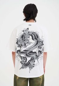 Witte katoen t-shirt met een groot zwart en grijs drakenafbeelding op de achterkant, omringd door bloemontwerpen en tekst.