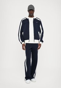 Training outfit bleumarin cu dungi albe pe laterale, jachetă cu fermoar și două buzunare frontale, purtată cu pantaloni asortați și adidași albi.