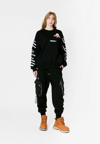 Sudadera negra con gráficos en blanco, que presenta un diseño floral y un bolsillo. Pantalones cargo negros con cremalleras y botas beige con suelas gruesas.