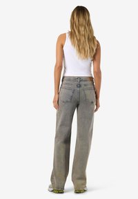 Sieviete ar gariem blondiem matiem, ģērbta baltā bezroku topā, izbalējušās taisnas džinsu biksēs un sporta apavos, stāvot ar muguru pret baltu fonu.