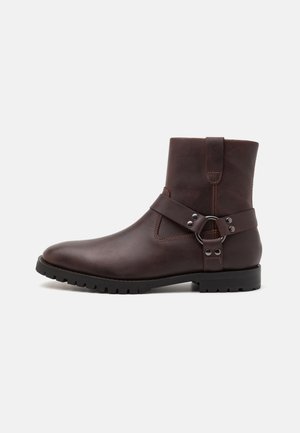 Brown leather ankle boot with a rounded toe, side strap, silver hardware accent, and rugged black outsole. Features smooth texture and minimal stitching.