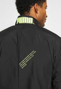 Svart jacka med hög krage som har en neon grön "PUMA"-logotyp. Textdetaljer i klar grön på baksidan framhäver hållbar produktion.