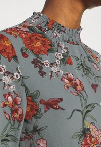 Primer plano de una persona que lleva una blusa gris de flores con cuello alto, con flores rojas, rosas y blancas, y un estampado de pájaros.