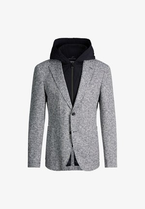 Blazer gris texturé porté par-dessus une veste à capuche noire zippée, combinant styles formel et décontracté.