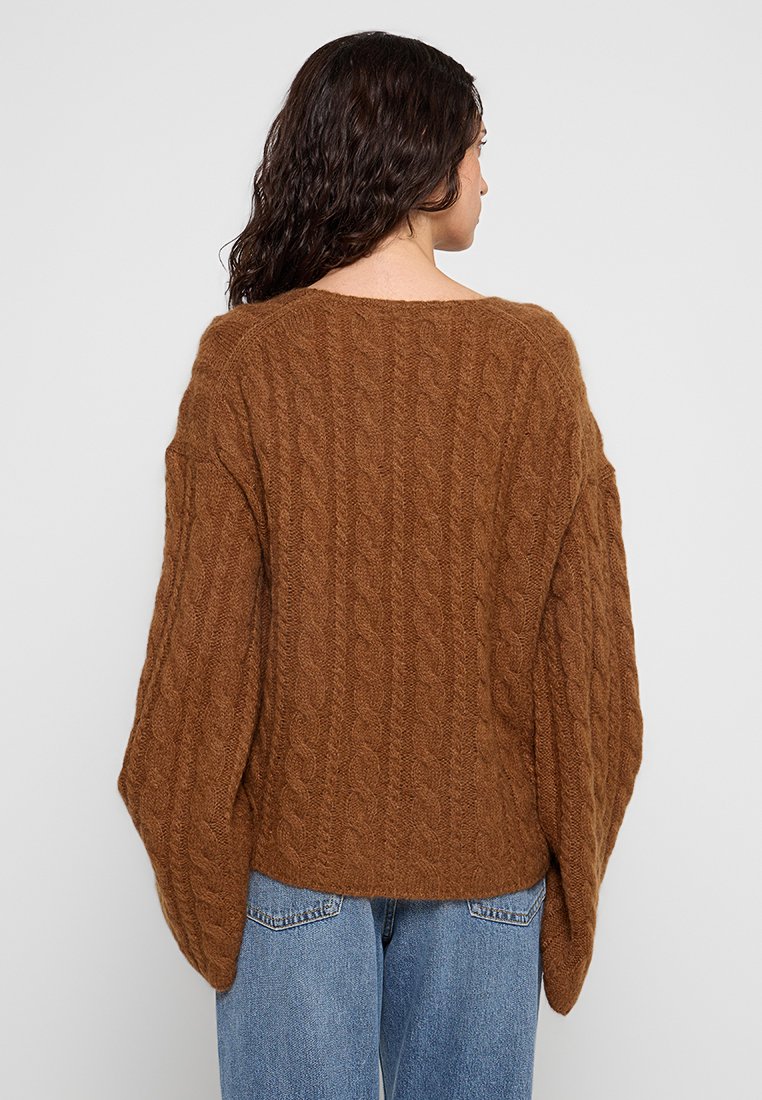 Brązowy sweter z dzianiny o splocie warkoczowym, o oversize'owym kroju, szerokich rękawach i okrągłym dekolcie. Tekstura wydaje się miękka i przytulna.