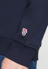 Sudadera azul marino con puños acanalados, que presenta un pequeño logotipo bordado en rojo y blanco en la manga izquierda. Fabricada en material de algodón suave.