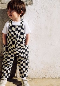 Salopette a quadretti in bianco e nero, in cotone, con grandi tasche, indossata sopra una t-shirt bianca dal taglio classico e scarpe da ginnastica leggere.