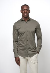 Camicia a manica lunga oliva in tessuto morbido, caratterizzata da una chiusura frontale con bottoni, bottoni neri e colletto classico. Abbinata a pantaloni beige chiaro.