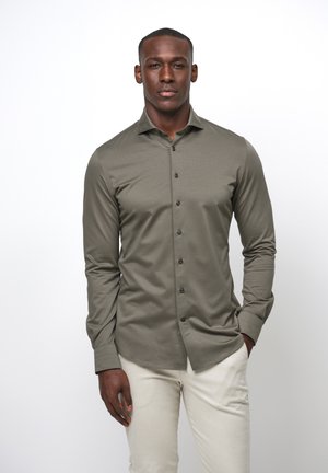 Olijfgroene blouse met lange mouwen van glad weefsel, met een knoopsluiting aan de voorkant, zwarte knopen en een klassieke kraag. Gecombineerd met lichtbeige broek.