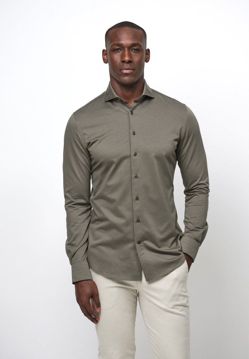 Camicia a manica lunga oliva in tessuto morbido, caratterizzata da una chiusura frontale con bottoni, bottoni neri e colletto classico. Abbinata a pantaloni beige chiaro.