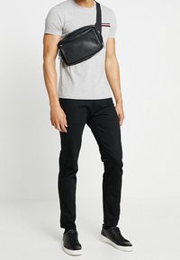 Schwarze Umhängetasche aus glattem Leder mit einem Reißverschluss und verstellbarem Gurt, getragen über einem grauen T-Shirt und schwarzen Jeans.
