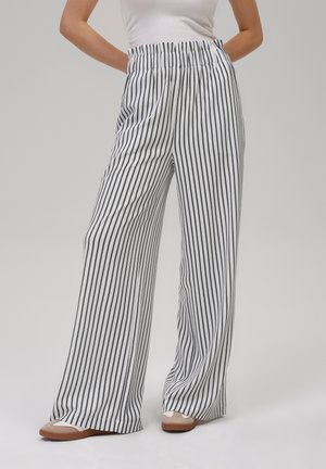 Weiße weitgeschnittene Hose mit schwarzen vertikalen Streifen. Mit elastischem Bund und fließender Textur. Kombiniert mit beigen Slippern.