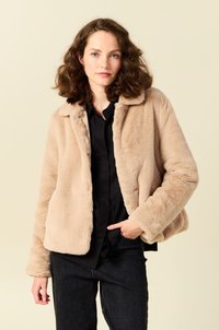 Beige nepbontjasje met kraag, lange mouwen en een ontspannen pasvorm. Draagt over een zwarte blouse, met een zachte, gestructureerde oppervlakte.