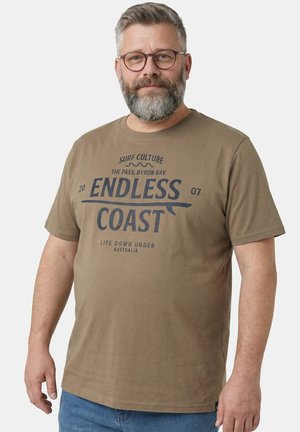 Brodaty mężczyzna w okularach, ubrany w brązowy t-shirt z napisem "Endless Coast" oraz niebieskie dżinsy, stojący na jasnym, jednolitym tle.