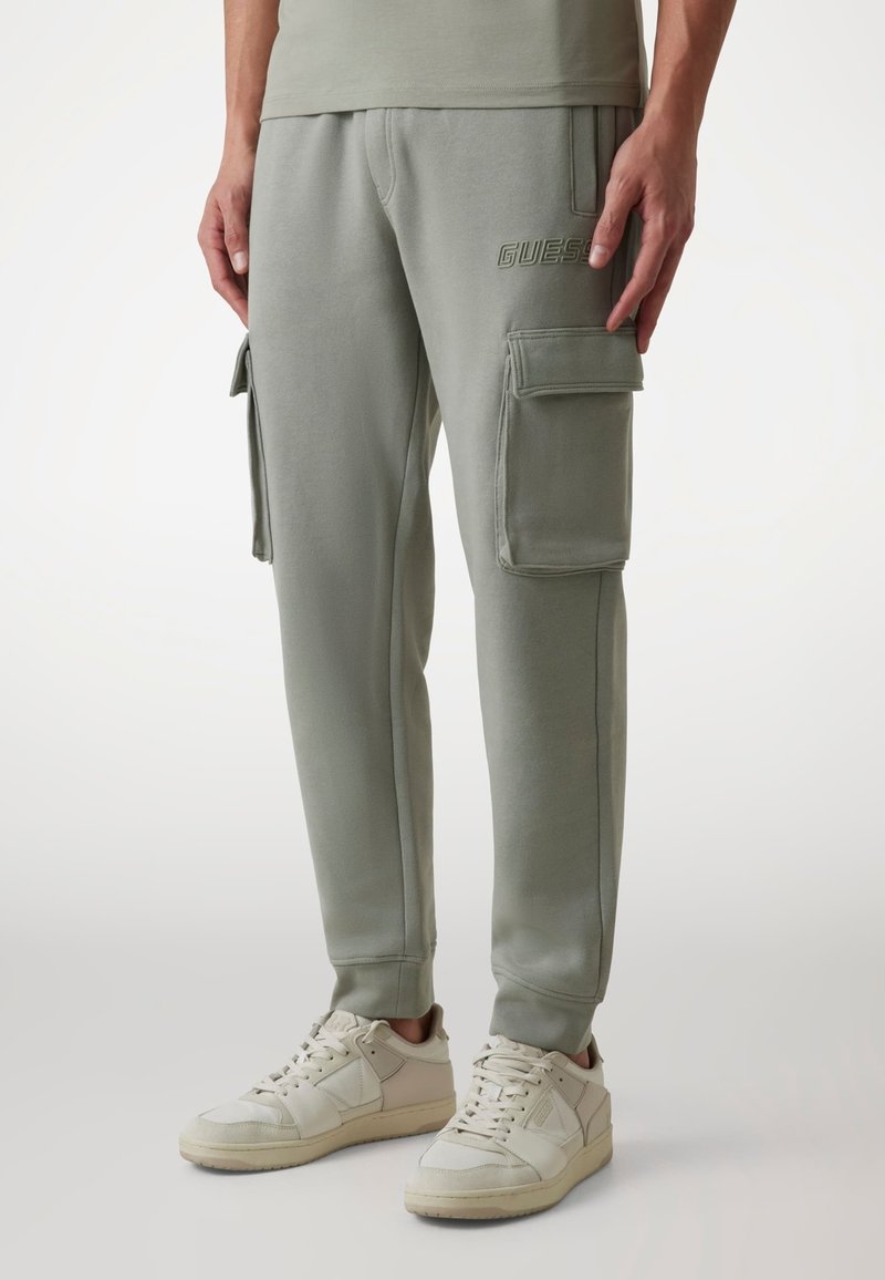 Jasnozielone spodnie cargo z dwoma bocznymi kieszeniami, miękką teksturą materiału, dopasowanym krojem i subtelnym logo "GUESS" z przodu. W połączeniu z białymi sneakersami.