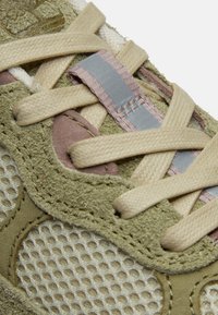 Gros plan sur une sneaker en daim vert et mesh avec des lacets beiges et une bande réfléchissante sur la languette.