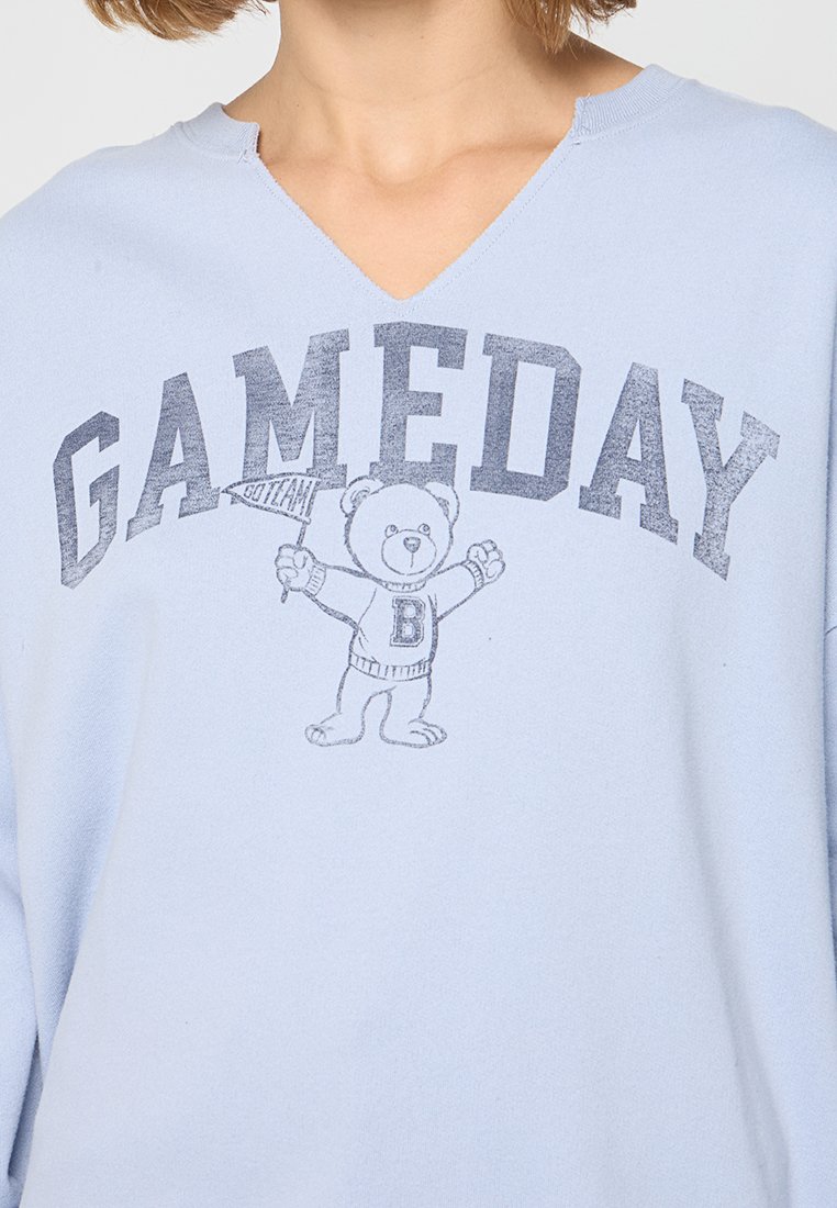 Helles blaues Sweatshirt mit V-Ausschnitt, bedrucktem "GAMEDAY"-Text und einer Illustration eines Bären, der ein Banner hält.