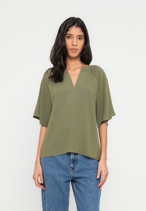 Olivgrüne Bluse mit V-Ausschnitt, kurzen weiten Ärmeln und strukturierter Stoff. Kombiniert mit hoch taillierten blauen Jeans.