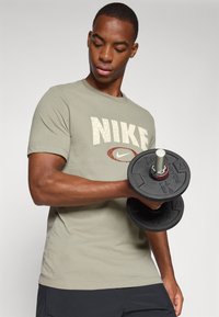Grågrønn Nike T-skjorte med en stor beige logo. Modellen holder en svart manual med fargede aksenter. Glatt stoff og tettsittende design.