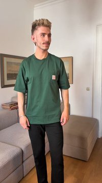 Žalias medvilninis marškinėliai su krūtinės kišene ir etikete, derinami su juodomis kelnėmis, ant medinio grindų paviršiaus šalia tekstūruotos šviesiai rudos sofos.