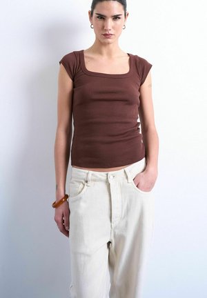 Woman with dark hair in a brown short-sleeve square-neck top and high-waisted white pants, posing with one hand in pocket against white wall.