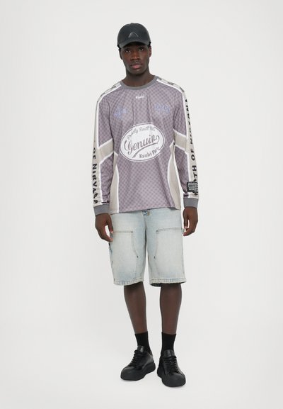 Grijze lange mouwen tshirt met grafische print, gecombineerd met lichtblauwe denim shorts. Het model draagt zwarte schoenen en een zwarte pet.
