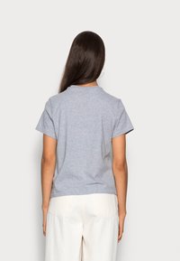 Nainen, jolla on pitkät tummat hiukset, pukeutuneena vaaleanharmaaseen lyhythihaiseen t-paitaan ja valkoisiin housuihin, seisoo kohtisuorasti vaaleata taustaa vasten.