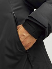 Chaqueta negra impermeable con un bolsillo con cremallera, tejido texturizado, puños reforzados y una silueta elegante y aerodinámica.