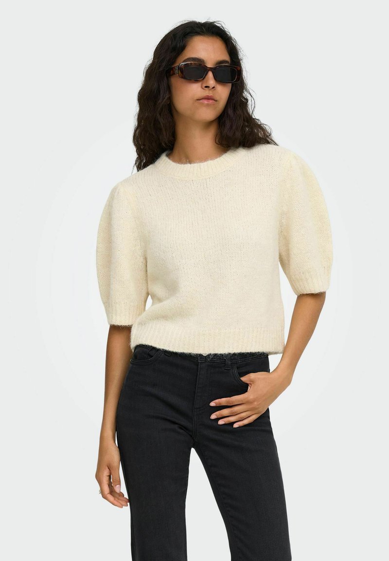 Femme portant un pull en maille crème à manches courtes, un jean noir et des lunettes de soleil rectangulaires, posant avec une main dans la poche devant un fond blanc.