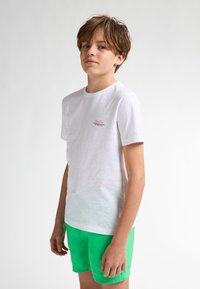 Bílá bavlněná tričko s krátkým rukávem a kulatým výstřihem, na hrudi má malé červené logo; doplněné zelenými plaveckými šortkami.