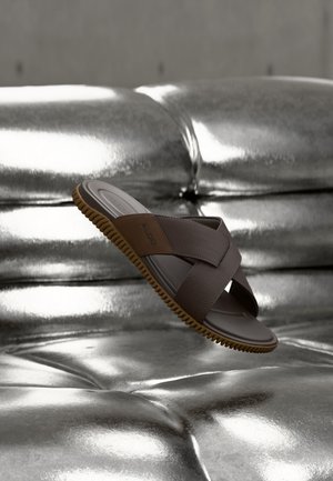 Brun ALDO skyvesandal med teksturerte remmer og riller på sålen som svever over en blank sølvfarget, tuftet overflate.