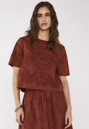 Donna con capelli mossi castano indossa un top e una gonna color ruggine con ricami floreali, abbinati a braccialetti e anelli dorati.