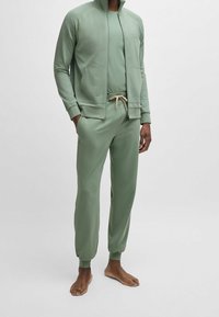 Groen trainingspak van zachte stof met een jack met rits, bijpassende broek en beige trekkoord. Heeft een aansluitend ontwerp en geribbelde manchetten.