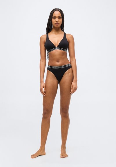 Femme aux cheveux tressés portant un soutien-gorge triangle noir Emporio Armani et une culotte assortie, debout pieds nus devant un fond clair uni.
