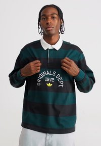 Pólo de mangas compridas, com riscas em verde escuro e preto. Apresenta um colarinho branco, logótipo com detalhes em amarelo e um corte descontraído.