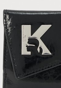 Čierna lesklá peňaženka z lakovanej kože, zdobená strieborným logom K s profilovým siluetom. Obsahuje dekoratívne švy.