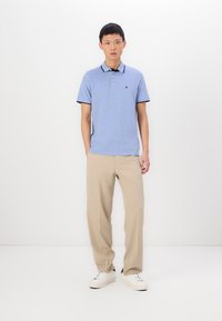 Light blue polo shirt with a black collar, short sleeves, and an embroidered logo. Paired with beige pants and white shoes.