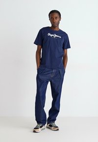 Tamsiai mėlynos spalvos medvilninė marškinė su "Pepe Jeans" logotipu, derinami su laisvais tamsiai džinsiniais kelnais ir daugiaspalviais sportbačiais.
