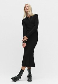 Musta pitkähihaisen neulemekko, jossa on vartalonmyötäinen malli, kaulus ja levenevä helma. Yhdistetty mustiin nilkkureihin, joissa on säädettävät soljet.