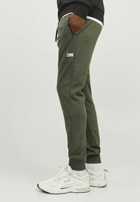 Olijfgroene joggingsbroek van zacht materiaal met een ritszak en een klein logopatch. Gecombineerd met witte hardloopschoenen.