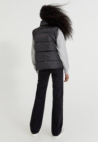 Czarna kamizelka puchowa z wysokim kołnierzem, z poziomymi szwami, noszona na szarym, długim rękawie oraz w komplecie z czarnymi spodniami.