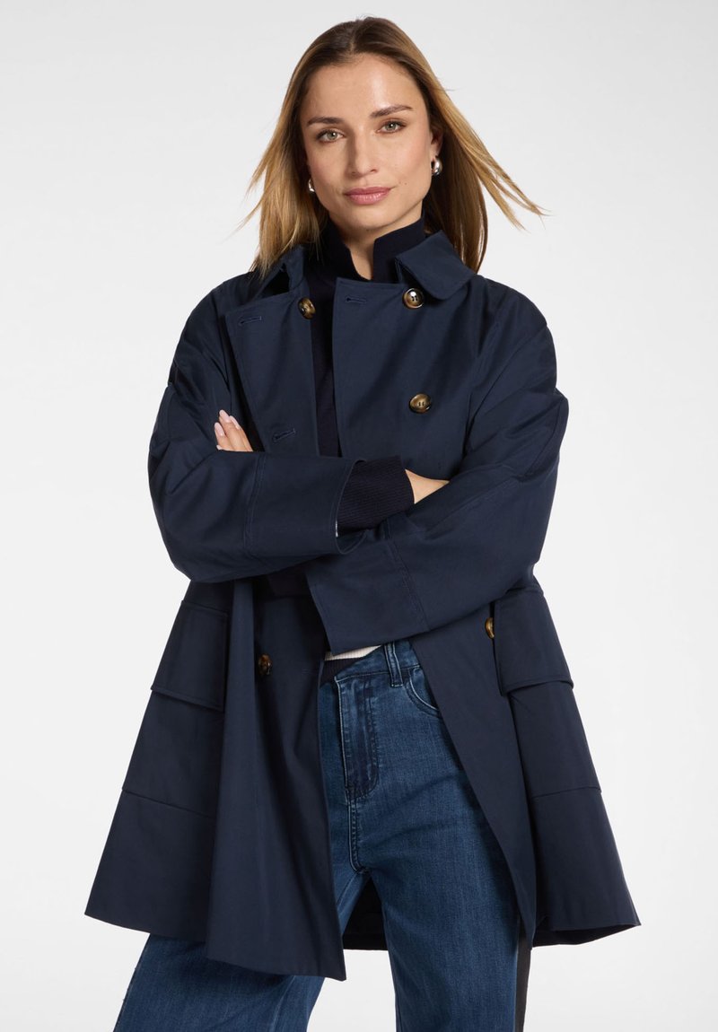 Donna con capelli biondi lisci che indossa un cappotto doppiopetto blu navy sopra un paio di jeans, in piedi con le braccia incrociate davanti a uno sfondo semplice.