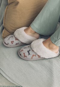 Graue Plüschpantoffeln mit Hundemotiven, ausgestattet mit weichem weißem Kunstpelzfutter, abgerundetem Zehenbereich und gesteppten Details. Getragen mit grüner Hose.