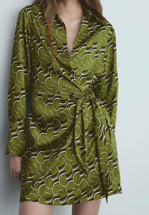 Mujer con vestido cruzado verde con un patrón abstracto curvo en blanco y negro, atado a la cintura, de pie frente a un fondo claro.