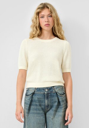 Jeune femme aux longs cheveux blonds ondulés portant un pull blanc texturé à manches courtes et un jean bleu taille haute.