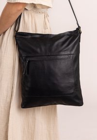 Black leather shoulder bag with a flat shape, features a front zip pocket, shoulder strap, and stitched detailing. Soft texture and minimalist design.