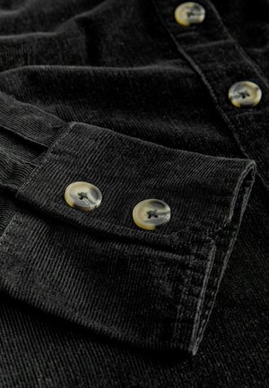 Mustmustekangast, millel on tekstuuriga viimistlus, millel on manseti peal kaks ümmargust tan värvi nööpi ja volditud krae.