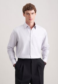 Camicia grigia chiara con taschino, maniche lunghe e colletto classico, abbinata a pantaloni neri con pieghe. Tessuto morbido.