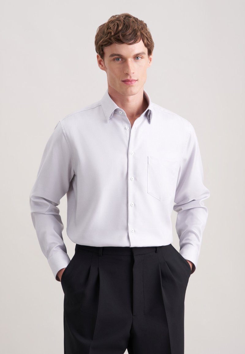 Camicia grigia chiara con taschino, maniche lunghe e colletto classico, abbinata a pantaloni neri con pieghe. Tessuto morbido.